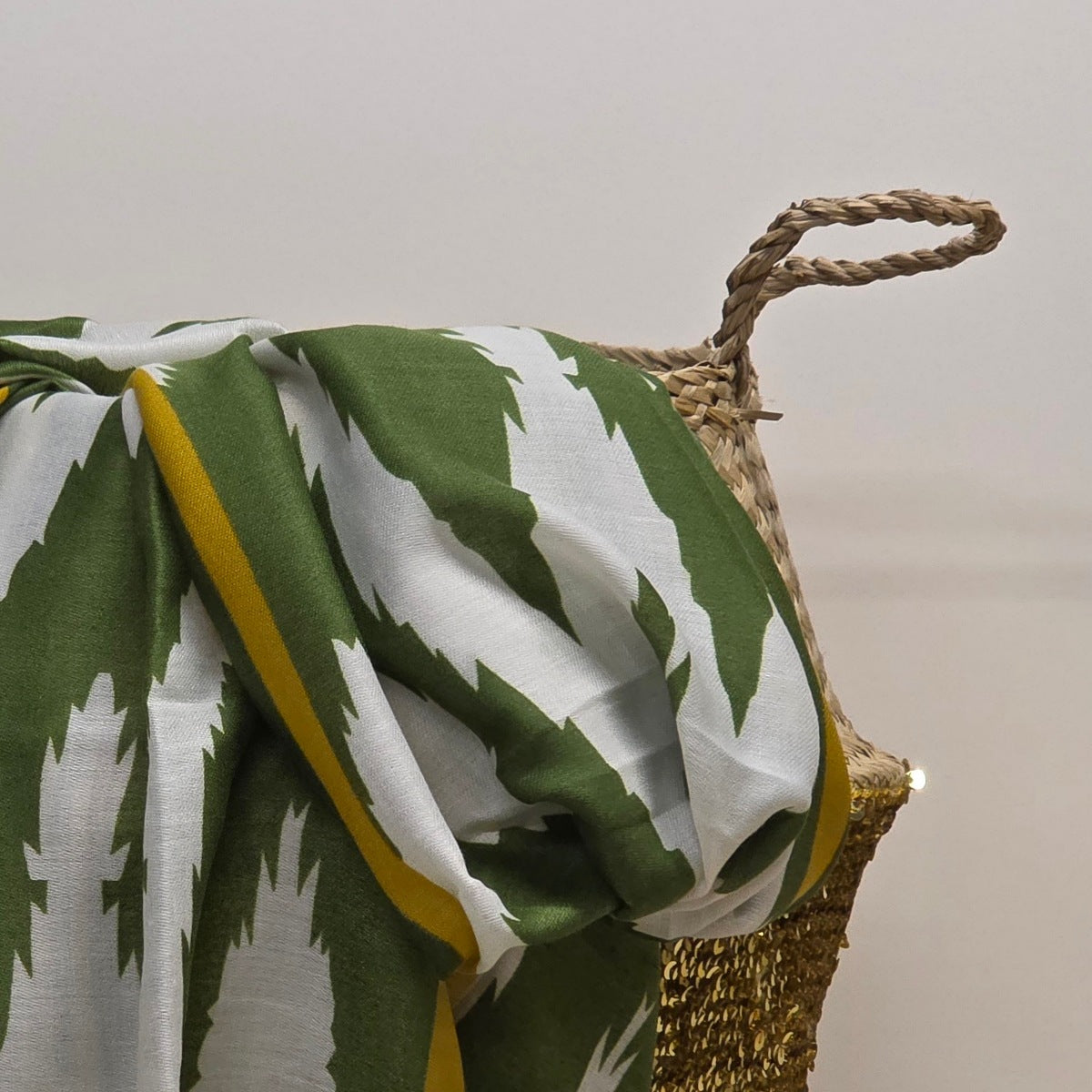 A green and white patterned throw blanket with a yellow border draped over a woven basket against a plain light background.