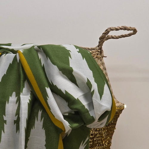 A green and white patterned throw blanket with a yellow border draped over a woven basket against a plain light background.
