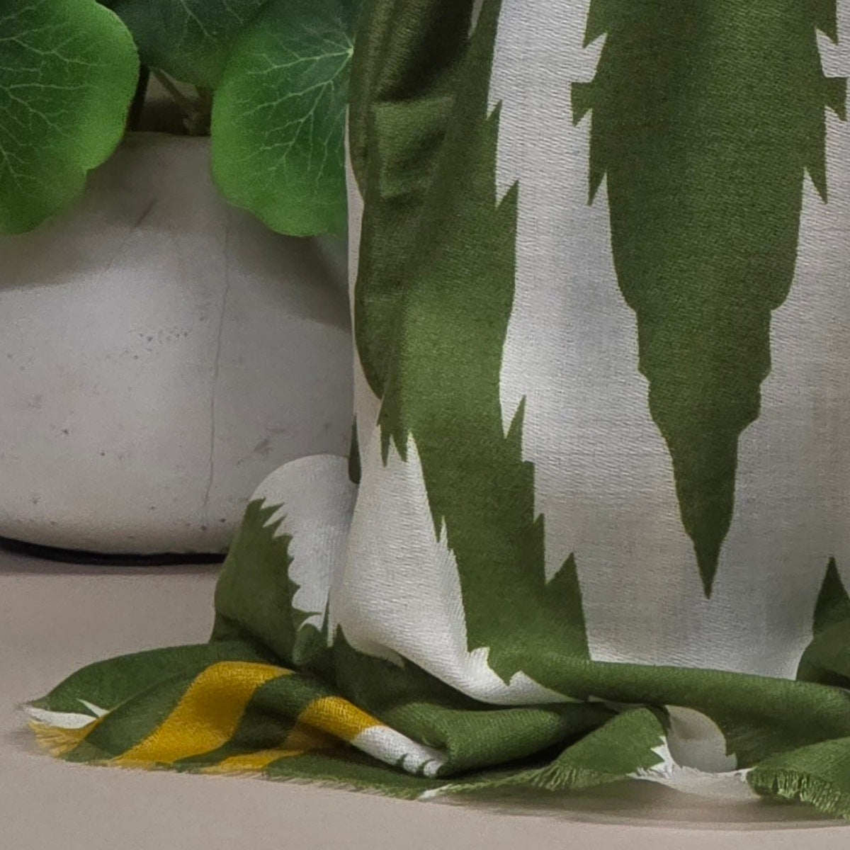 A close-up of a draped fabric scarf with a bold green and white jagged vertical pattern, with a yellow stripe visible near the lower left edge. The fabric is styled next to a white planter with green leaves.