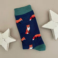 A single navy blue sock with an all-over pattern of red and orange foxes.  Green ribbed cuffs and heels, styled on a light beige surface with white star props.
