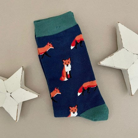 A single navy blue sock with an all-over pattern of red and orange foxes.  Green ribbed cuffs and heels, styled on a light beige surface with white star props.
