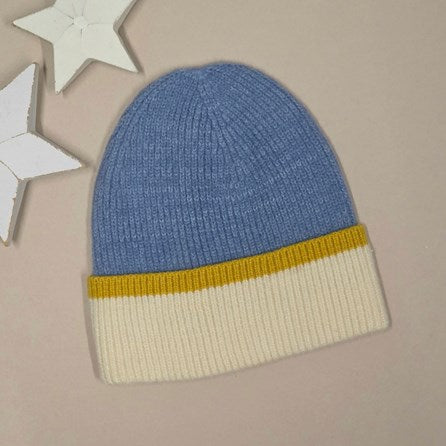 A ribbed knit beanie hat in mid blue with a mustard yellow band and an off-white cuff, displayed on a neutral grey background with white star-shaped decorative props.