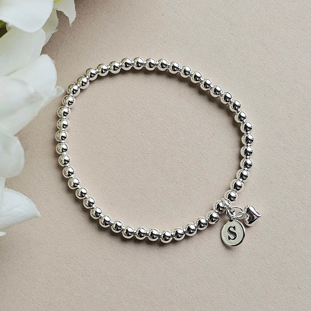 A silver beaded bracelet with a small round initial 'S' charm and a tiny bell charm, displayed on a light beige surface with white flowers in the corner.